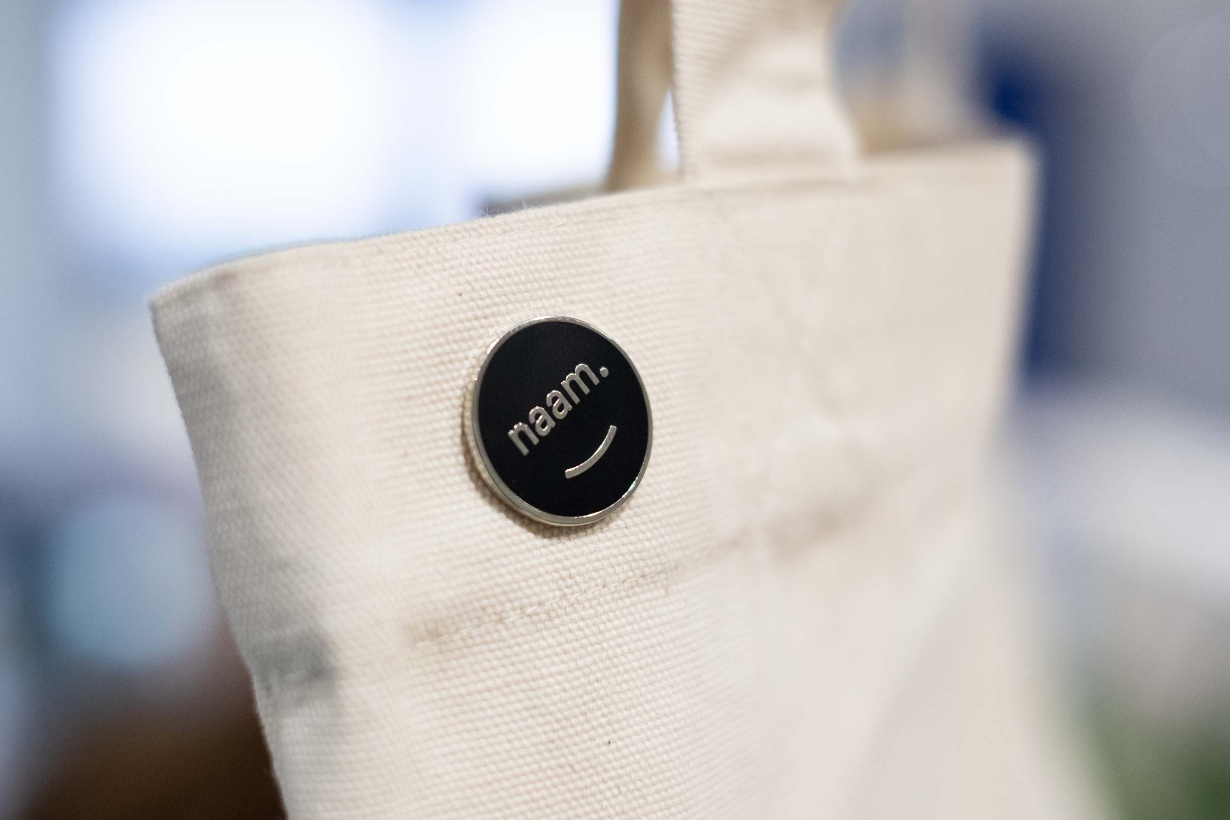 A canvas tote bag with a Naam logo enamel pin